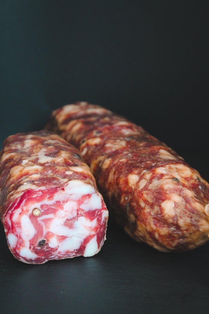 Two rustic cured salami sticks with marbled texture on a dark surface, perfect for meat lovers.